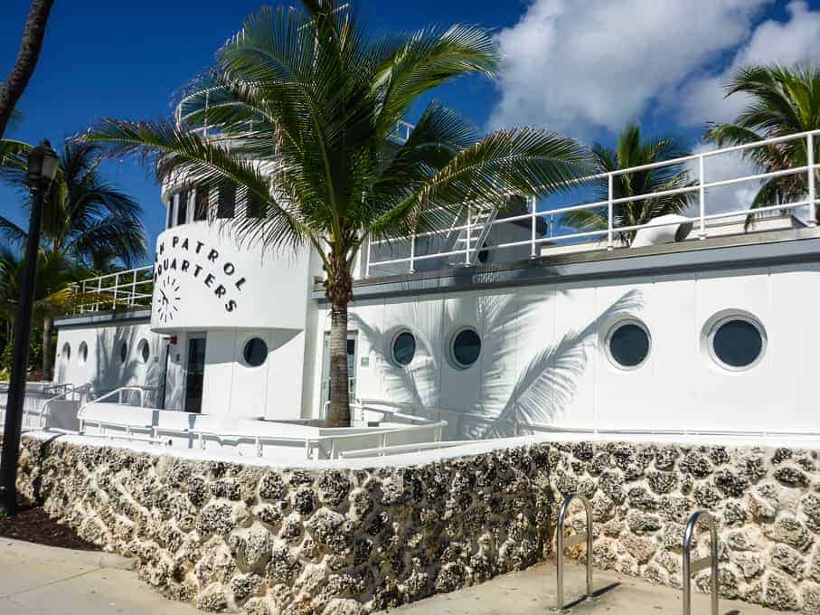 White Art Deco building labeled "Patrol Quarters" with palm trees and a stone wall in front, under a blue sky.