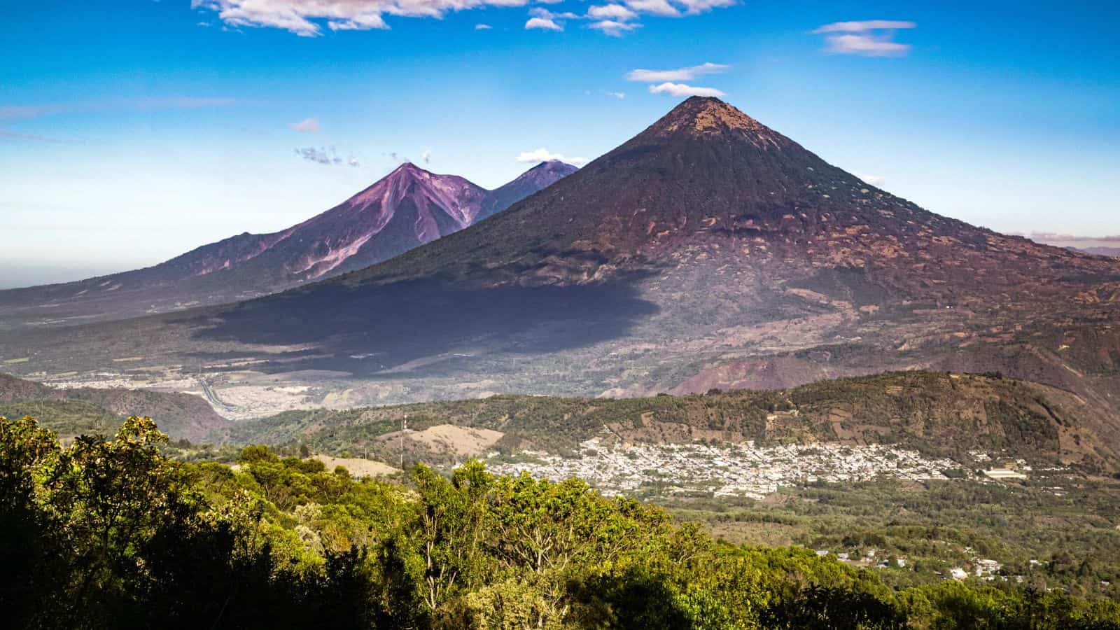A wide view of two large volcanoes with conical peaks, surrounded by green landscape and a small town at the base, under a clear blue sky with a few clouds.