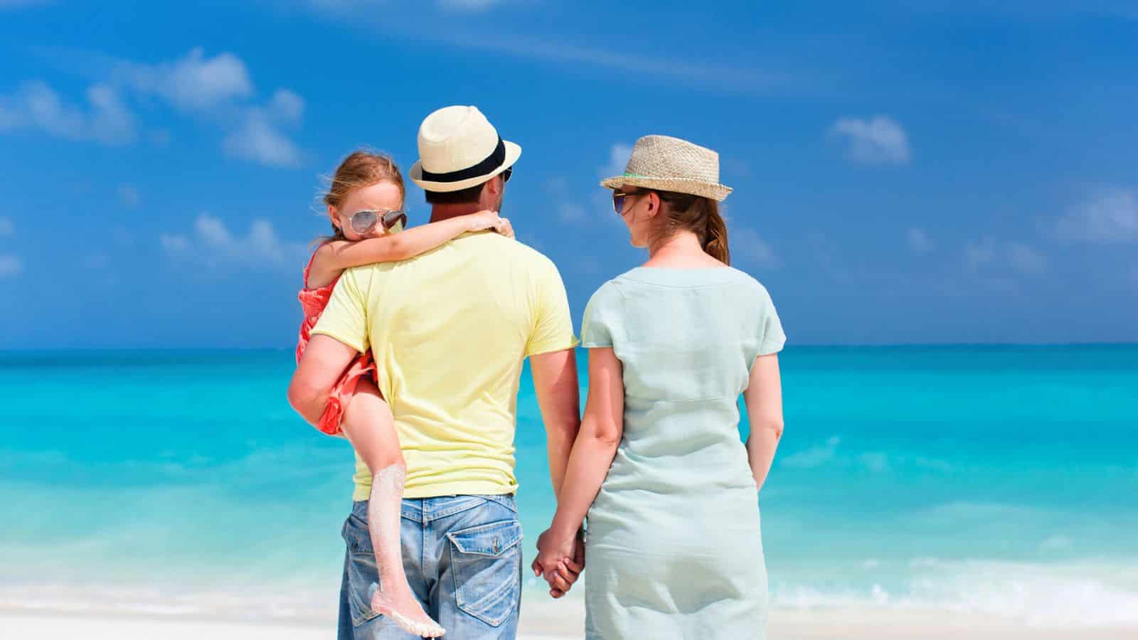 Family on a beach: man carries a child, woman holds his hand, all facing the ocean under a blue sky.