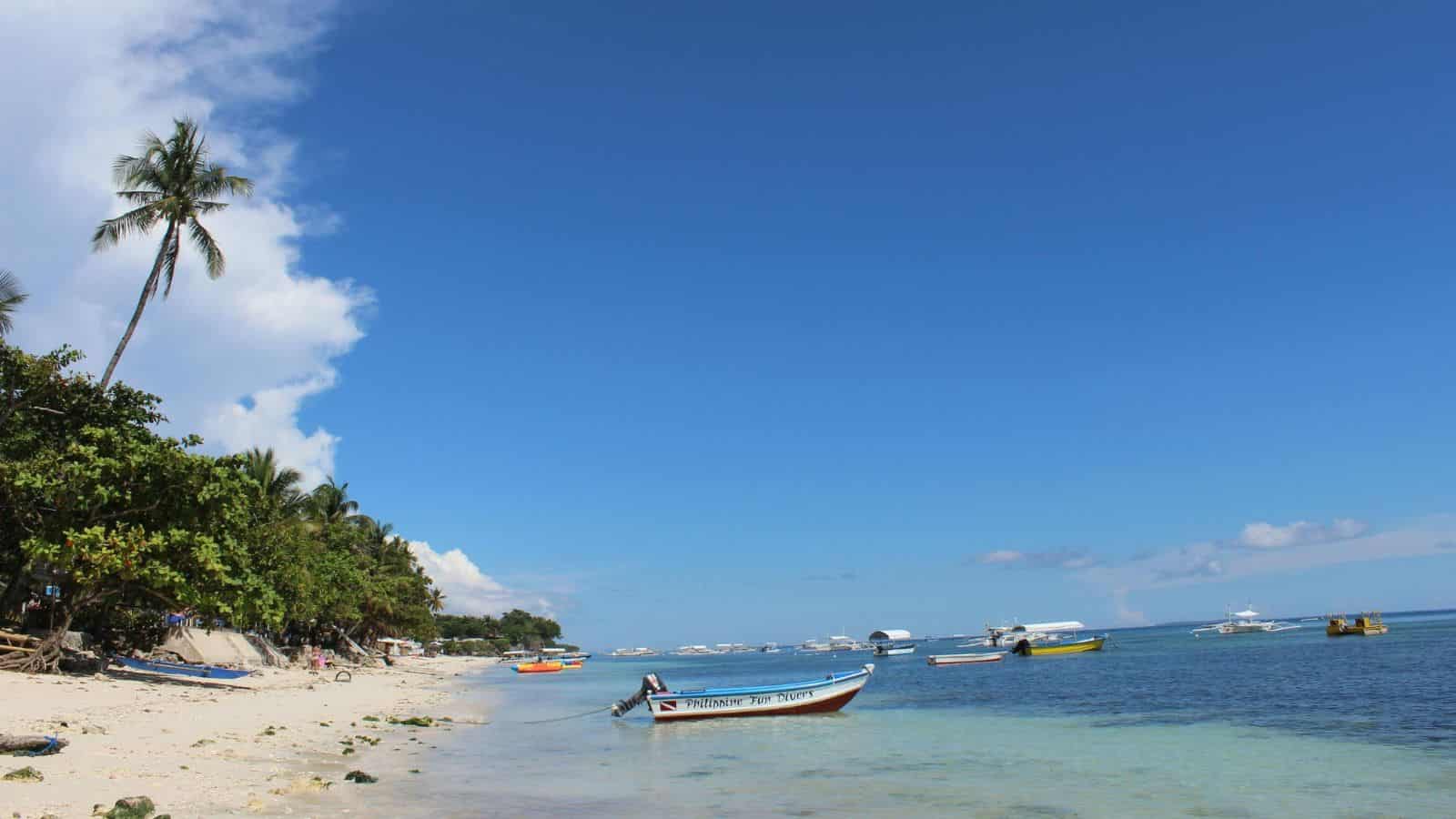 Tropical beach with boats on clear blue water, palm trees, and a bright blue sky with few clouds.