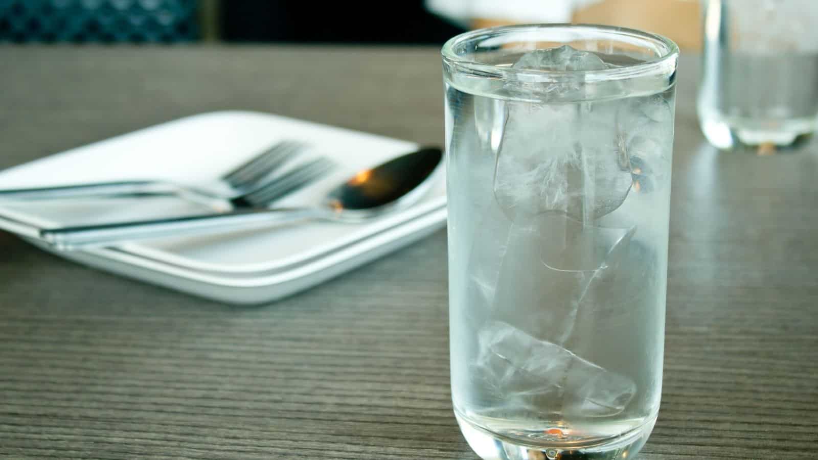 A clear glass filled with ice water sits on a wooden table, with a stack of white plates and a fork and spoon in the background.