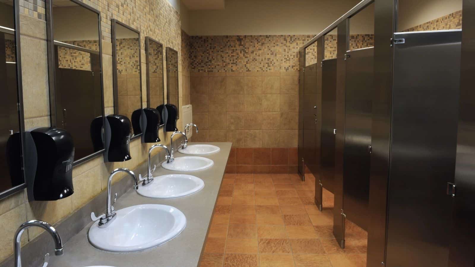 A public restroom with a row of five sinks, mirrors, and soap dispensers on the left, and several closed metal stall doors on the right—designed in neutral tones, it may cause travel confusion for first-time travelers adjusting to European habits.