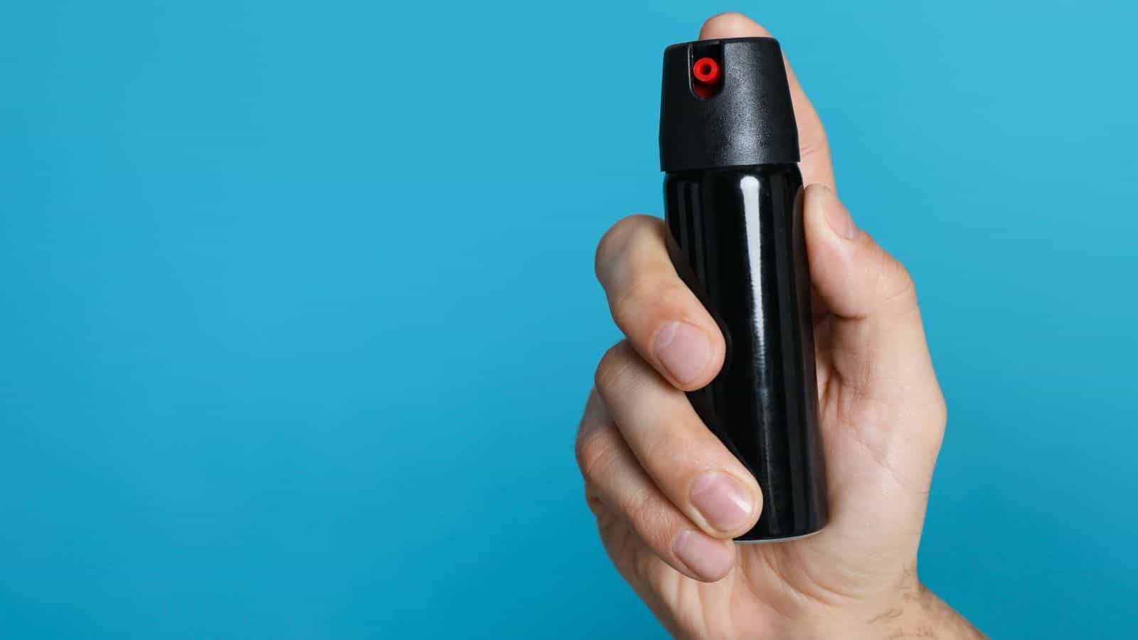 A hand holds a small black aerosol spray can with a red nozzle&mdash;one of the most common confiscated items by TSA agents&mdash;against a solid blue background, highlighting travel security concerns.