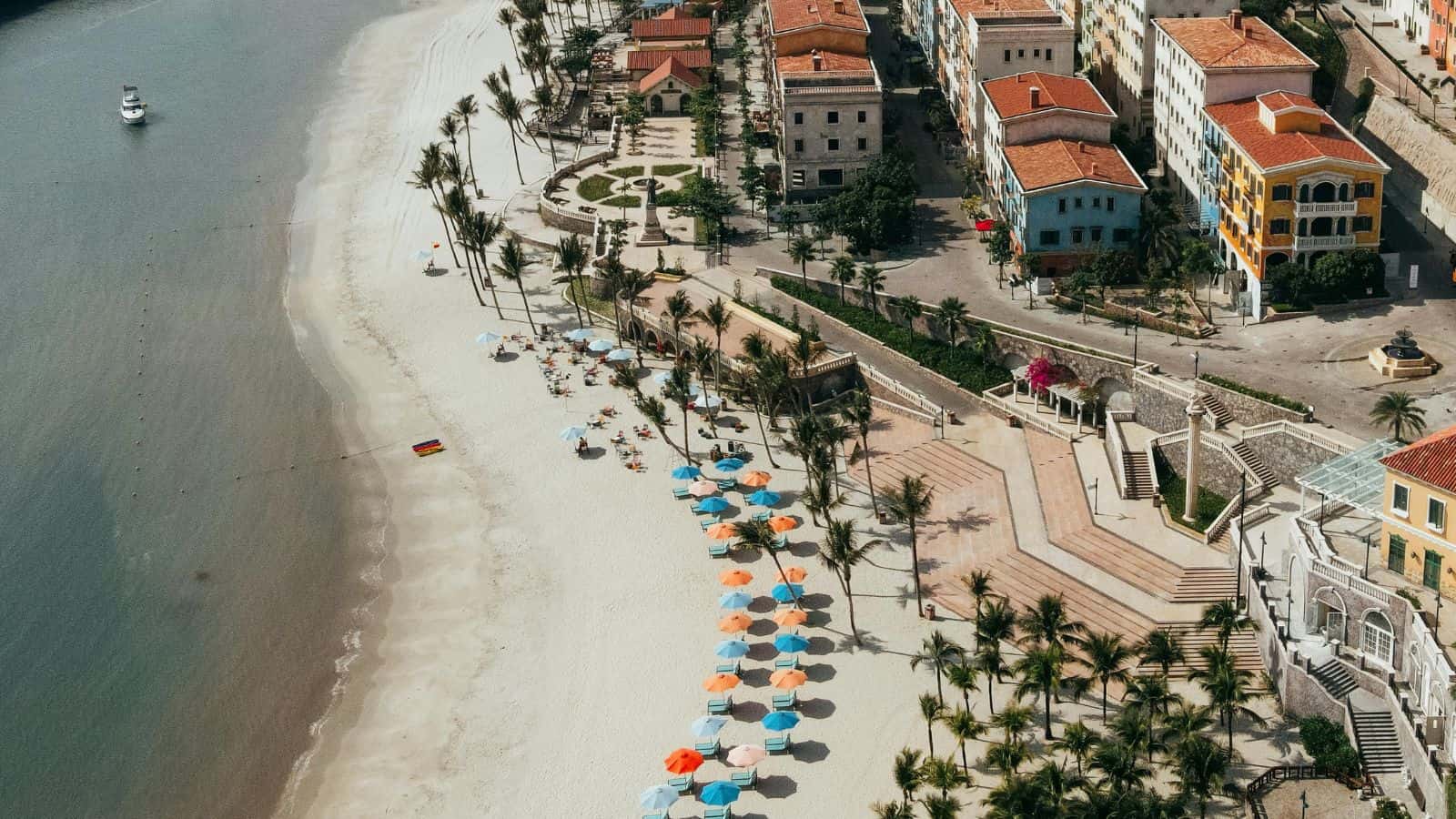 Aerial view of a beach lined with colorful umbrellas, palm trees, and nearby buildings with red roofs.