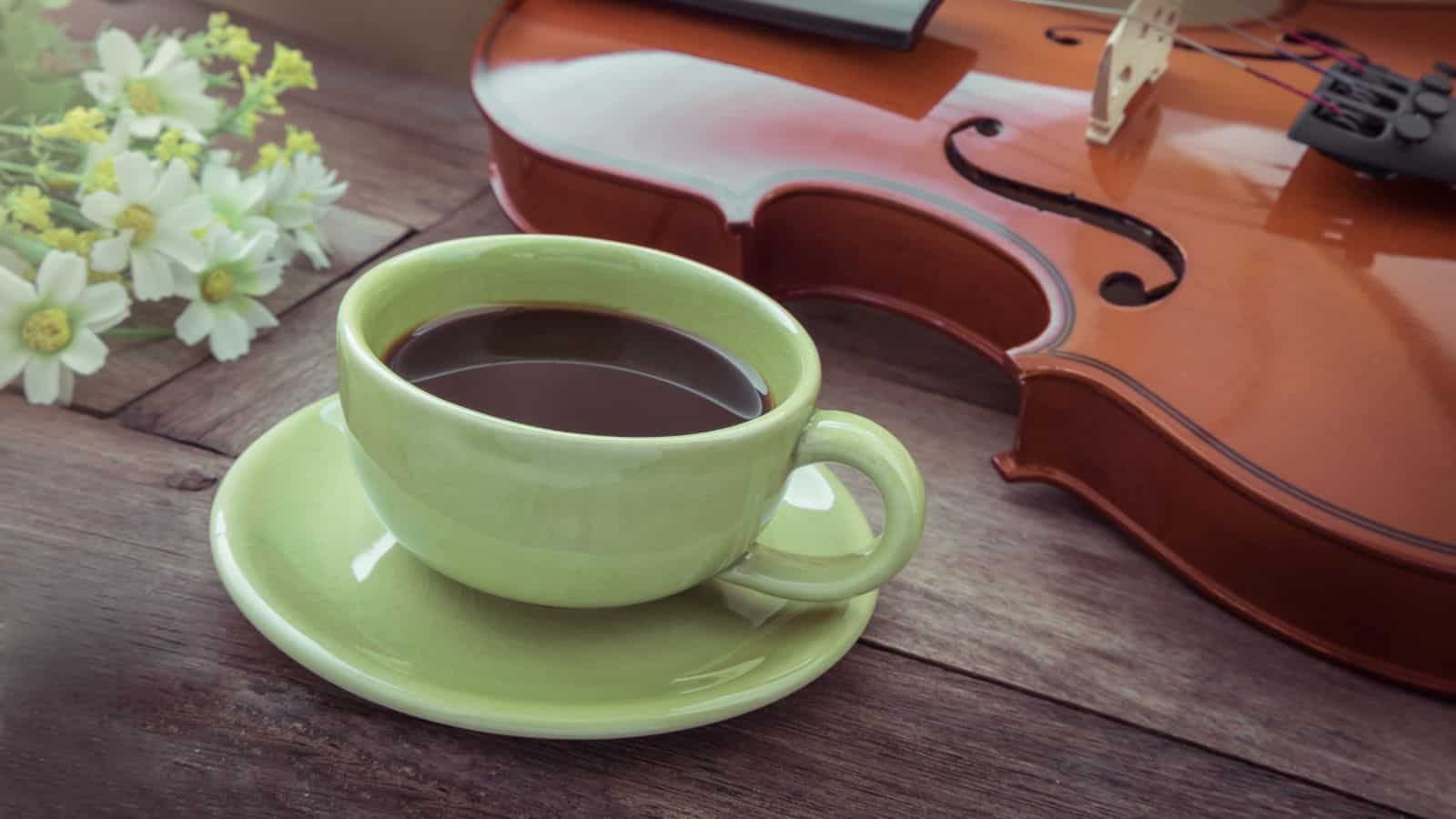 A green cup and saucer filled with black coffee sit on a wooden table next to white flowers and a violin placed on its side.
