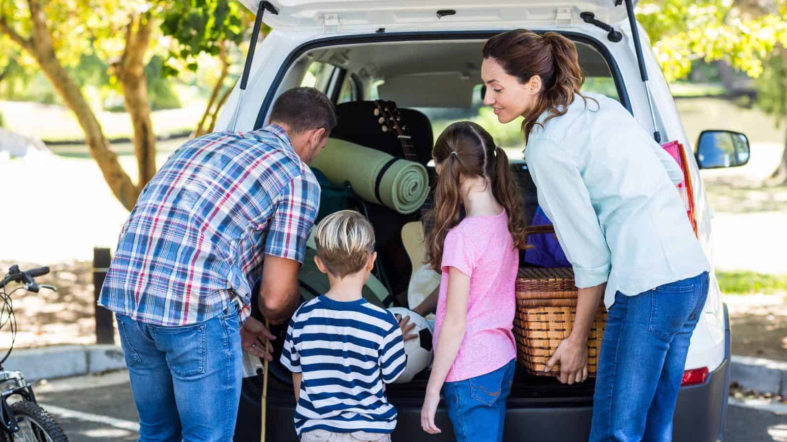 A man, woman, and two children stand at the open trunk of a car outdoors, loading items such as a basket and rolled mats, preparing for an outing or picnic.