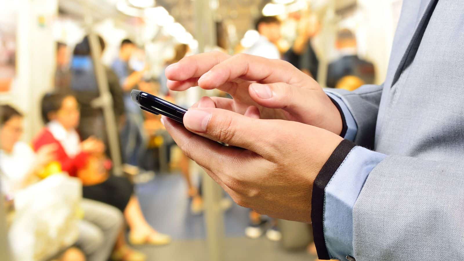 A person in a light blue suit uses a smartphone during a busy train ride, standing among other passengers on the crowded subway with blurred figures in the background.