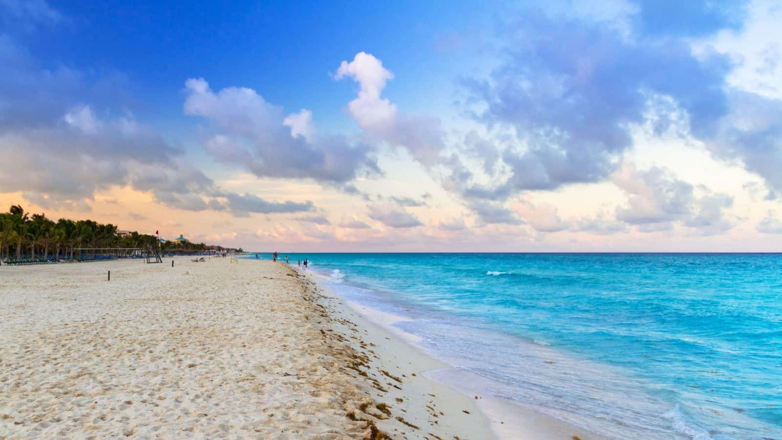 Wide sandy beach with gentle waves along clear turquoise water. Few people walk near the shoreline, and a line of trees is visible on the left. The sky is partly cloudy with soft light from the setting sun.