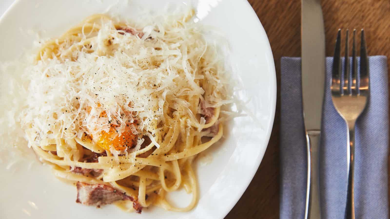 A plate of pasta topped with grated cheese and black pepper sits on a wooden table, making you jealous of those classic U.S. things that other countries wish they had, like this perfectly set fork and knife on a folded blue napkin.