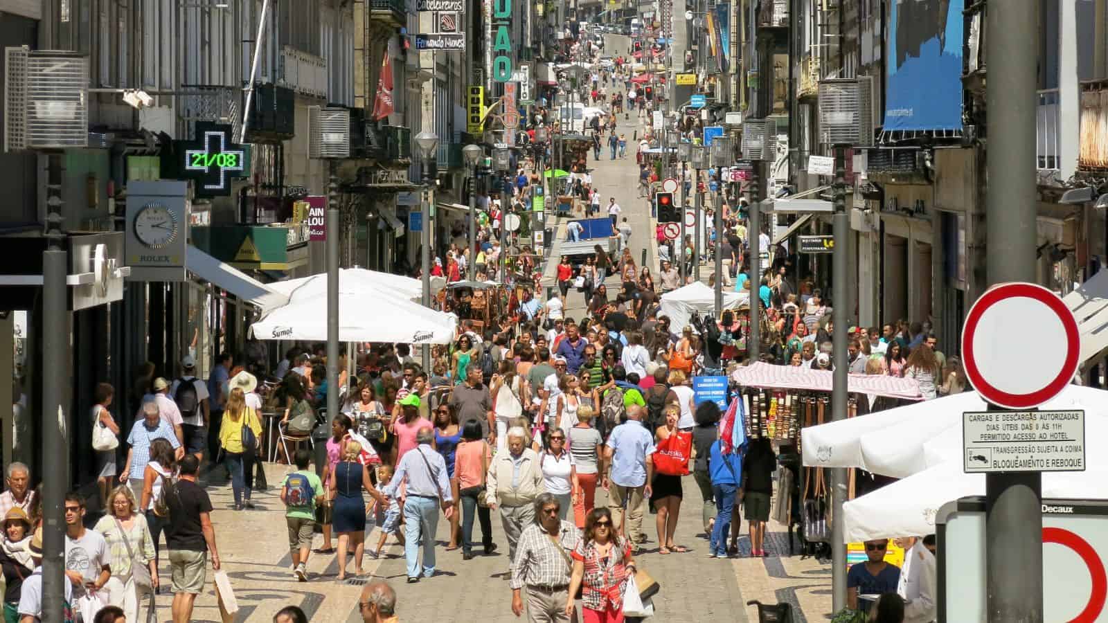 Crowded pedestrian street with shops, cafes, and people walking, shopping, and socializing on a sunny day.