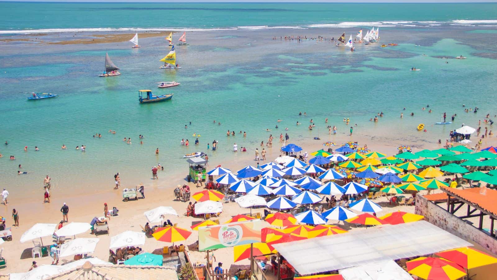 A crowded beach scene with many people swimming in clear turquoise water and numerous colorful umbrellas and beach chairs covering the sandy shore. Several boats and sailboats are visible in the water.