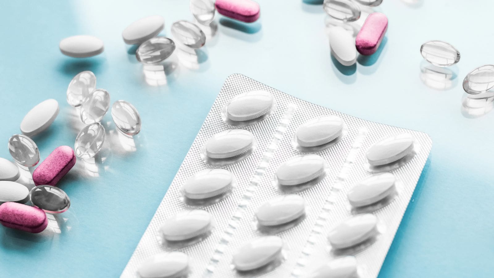 A close-up of various pills and capsules, including a blister pack of white tablets, loose pink and white capsules, and clear gel capsules scattered on a light blue surface.