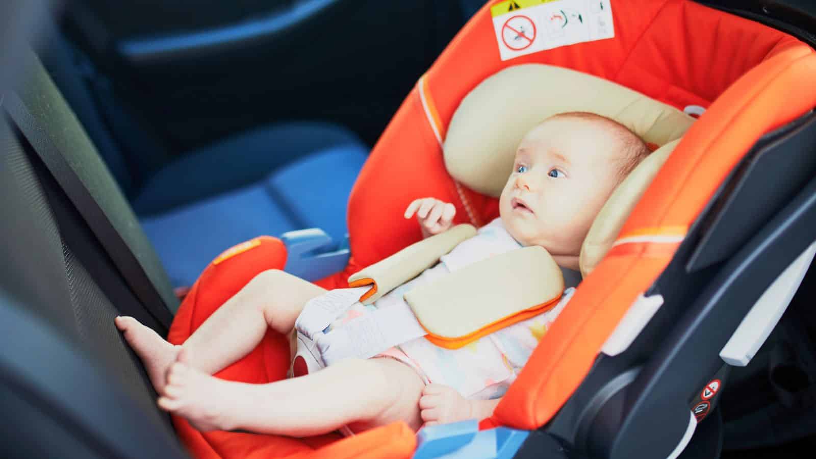 A baby in an orange car seat, securely fastened and facing backwards in a car.