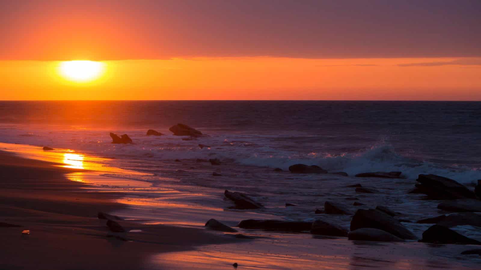 Sunset over the ocean with waves crashing on a rocky beach and colorful reflections on the wet sand.