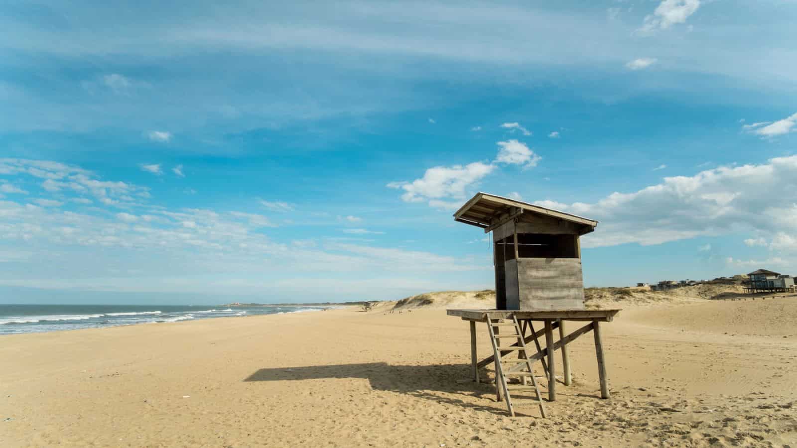 A wooden lifeguard tower stands on a deserted sandy beach under a blue sky with scattered clouds.