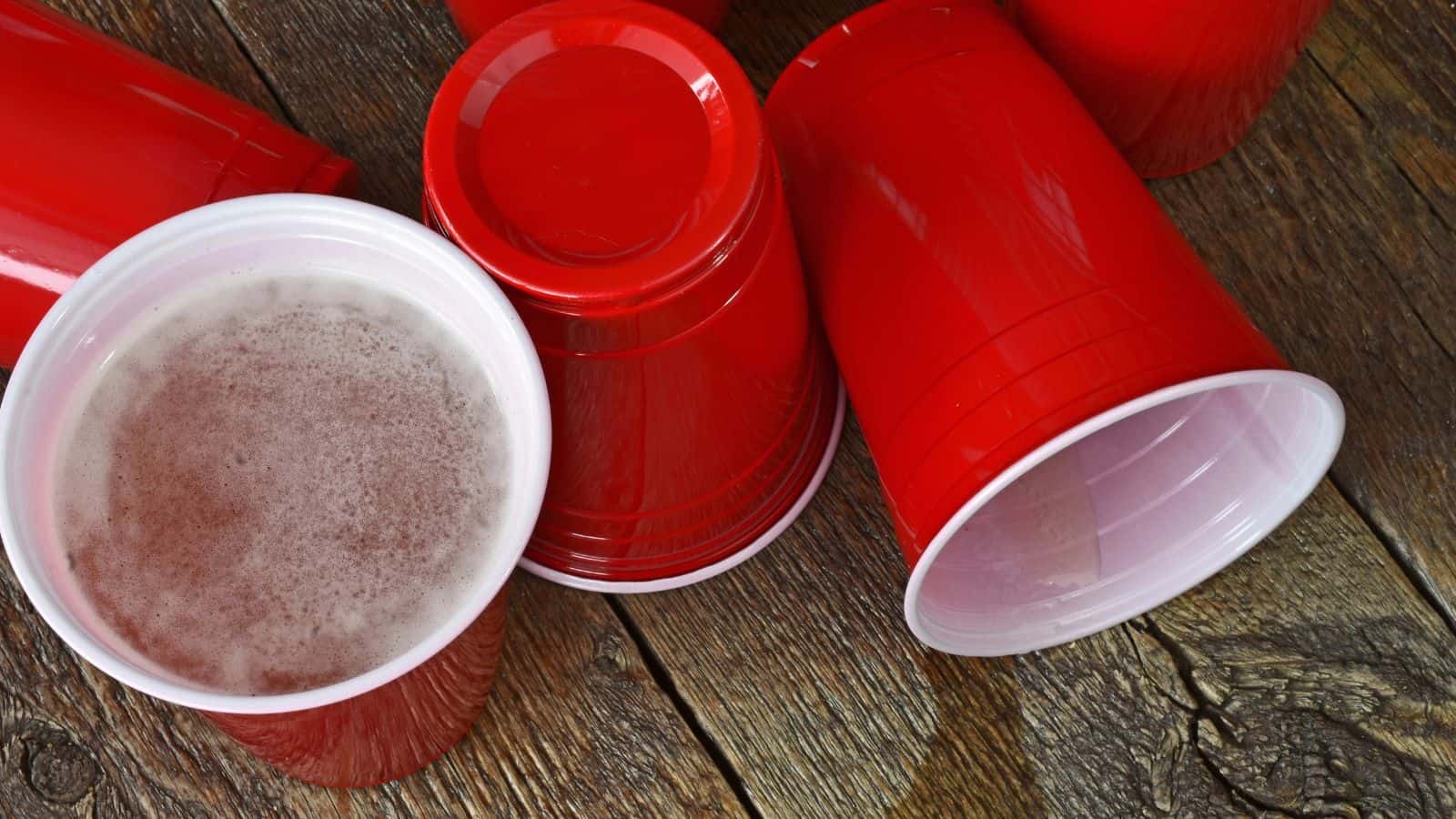 Several red plastic cups are on a wooden surface; one cup is upright and filled with a foamy beverage, while the others are empty and lying on their sides.