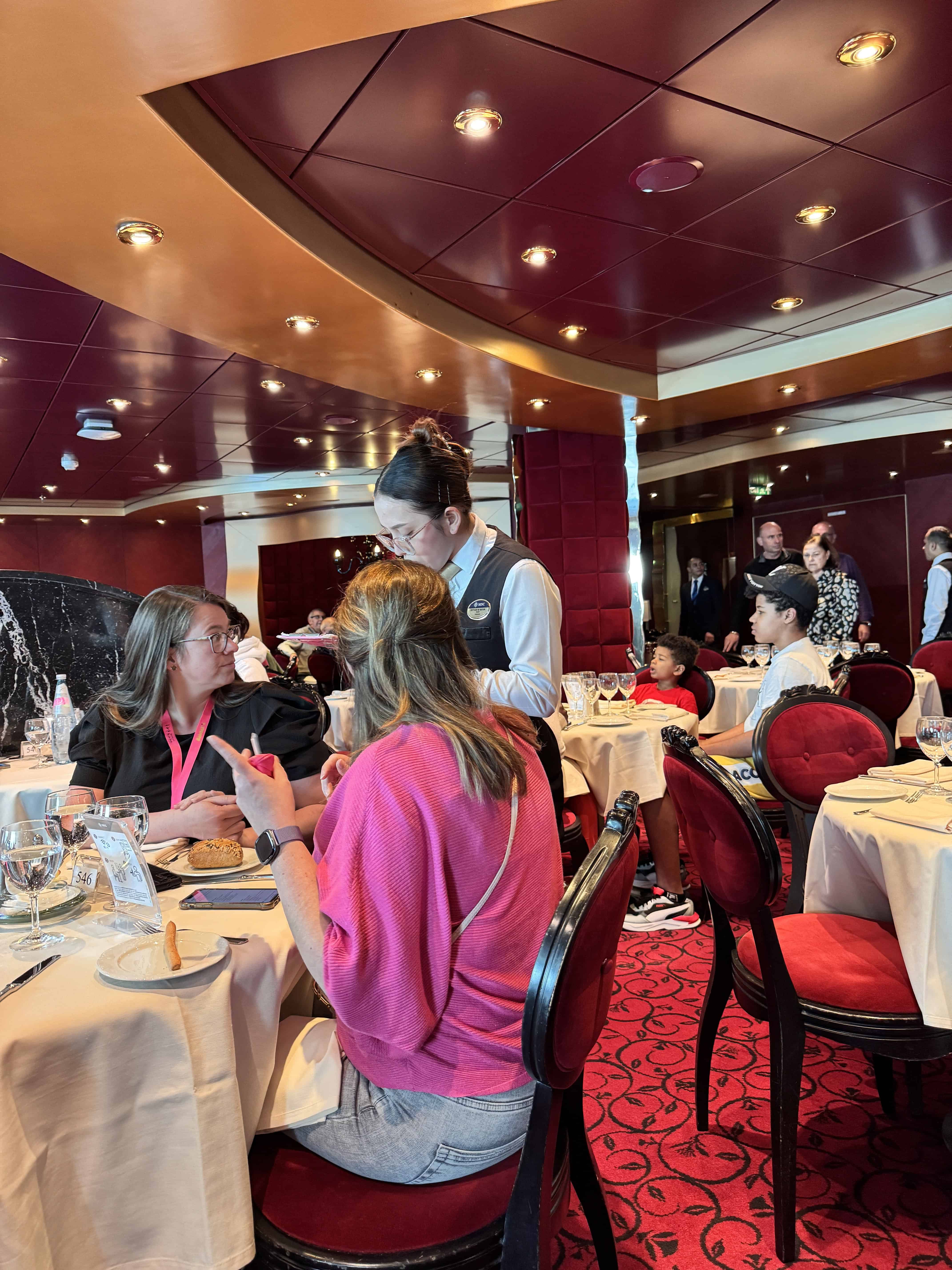A server takes an order at a lively restaurant with red decor and diners seated at round tables.