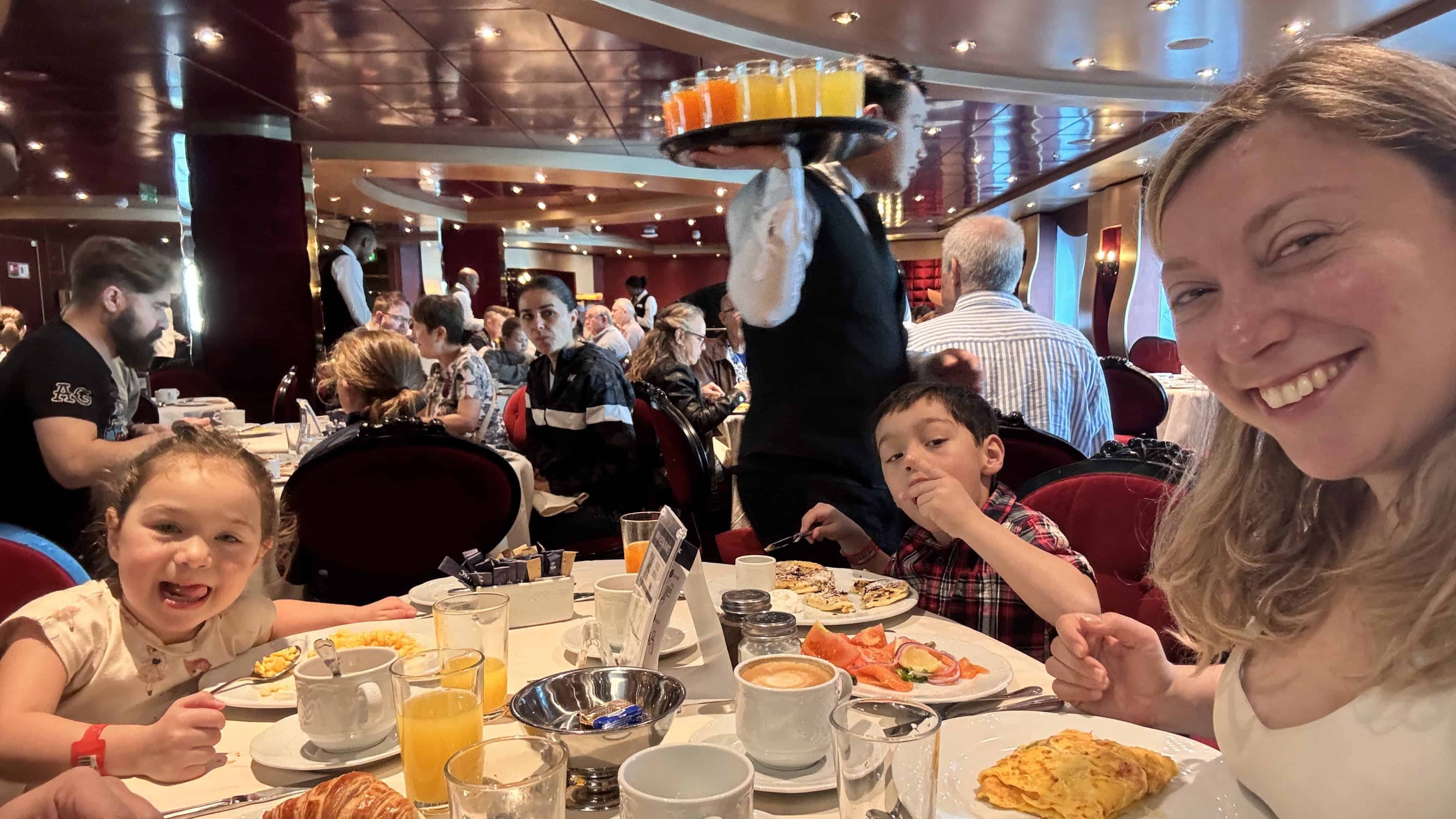 A woman and two children smile while having breakfast in a busy restaurant aboard MSC Fantasia Cruise, enjoying a memorable family cruise experience as a server carries drinks.