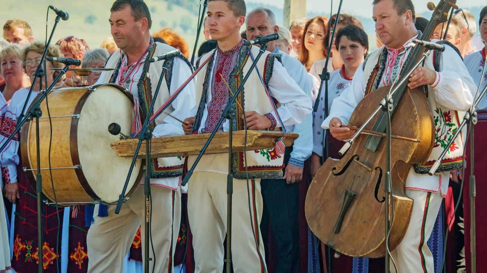 Musicians in traditional folk costumes play instruments at an outdoor cultural event with a crowd behind them.