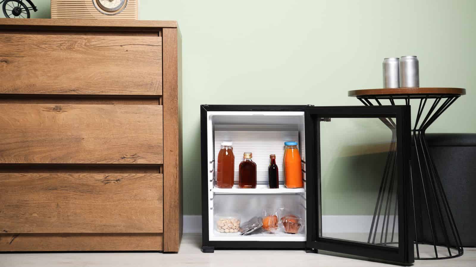 A small fridge with drinks and snacks sits open next to a wooden dresser and a round side table.