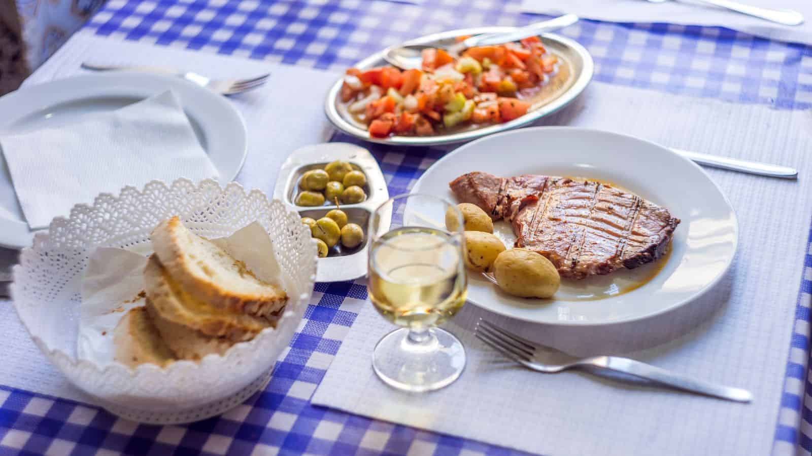 A plate with steak, potatoes, salad, bread, olives, and a glass of white wine on a checkered tablecloth.