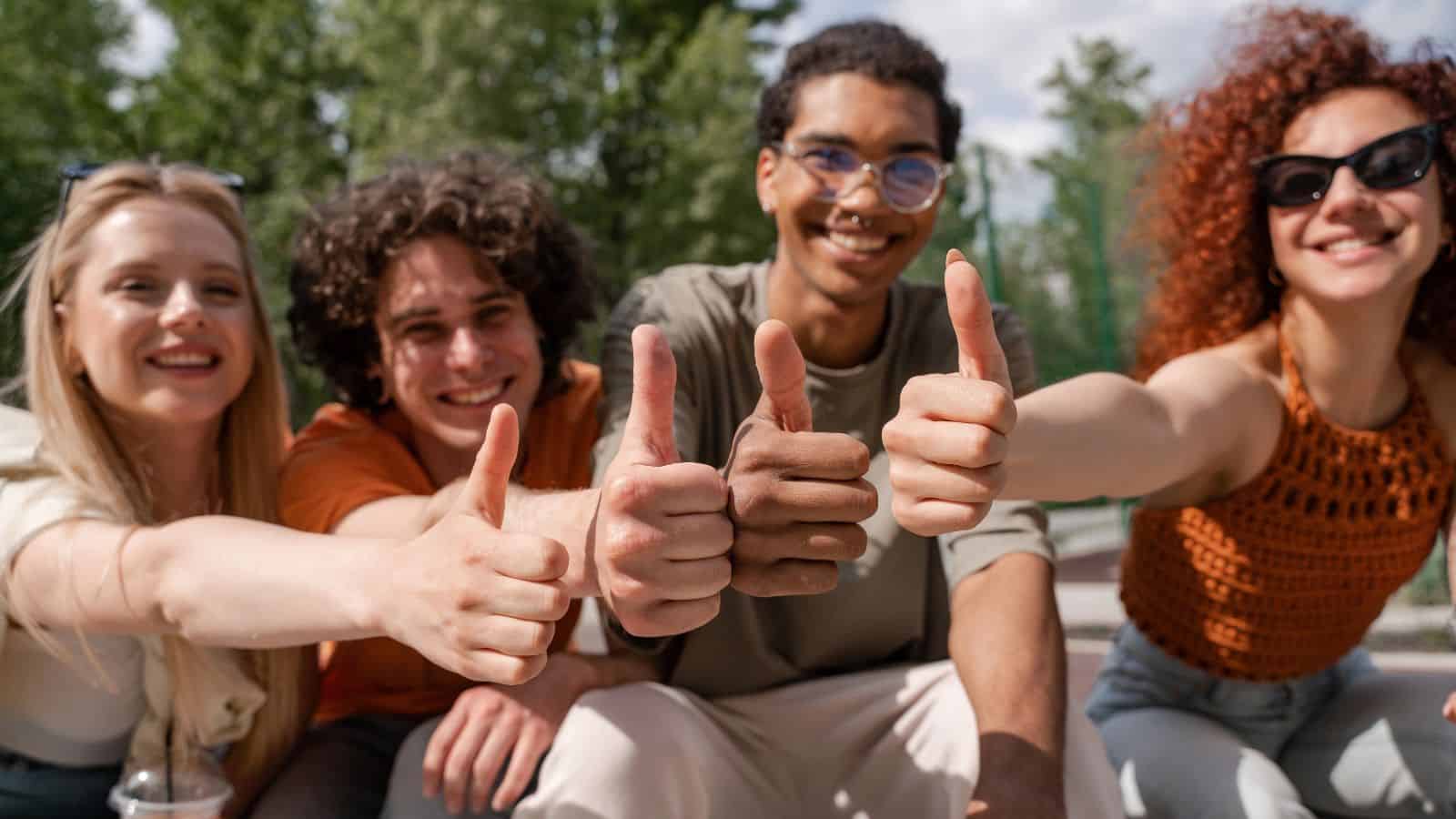 Four young adults sitting outdoors, smiling, and giving a thumbs-up gesture toward the camera. Trees and a bright sky are visible in the background.