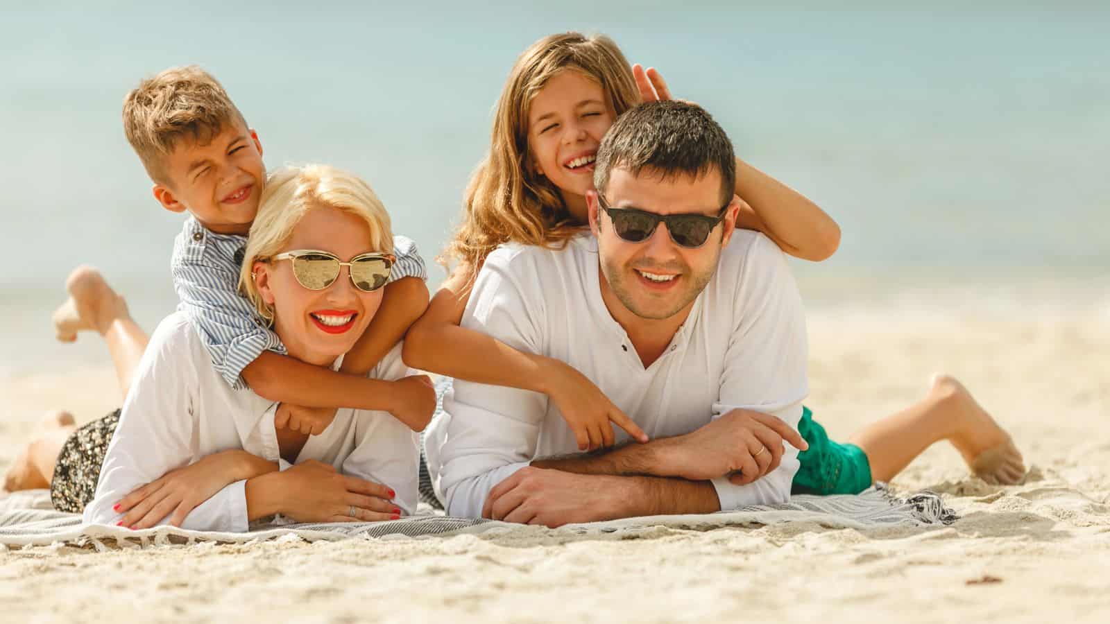 A man, woman, boy, and girl lie on a beach towel at the seaside. The adults and girl wear sunglasses. Everyone is smiling, and the boy and girl are playfully hugging the adults from behind.