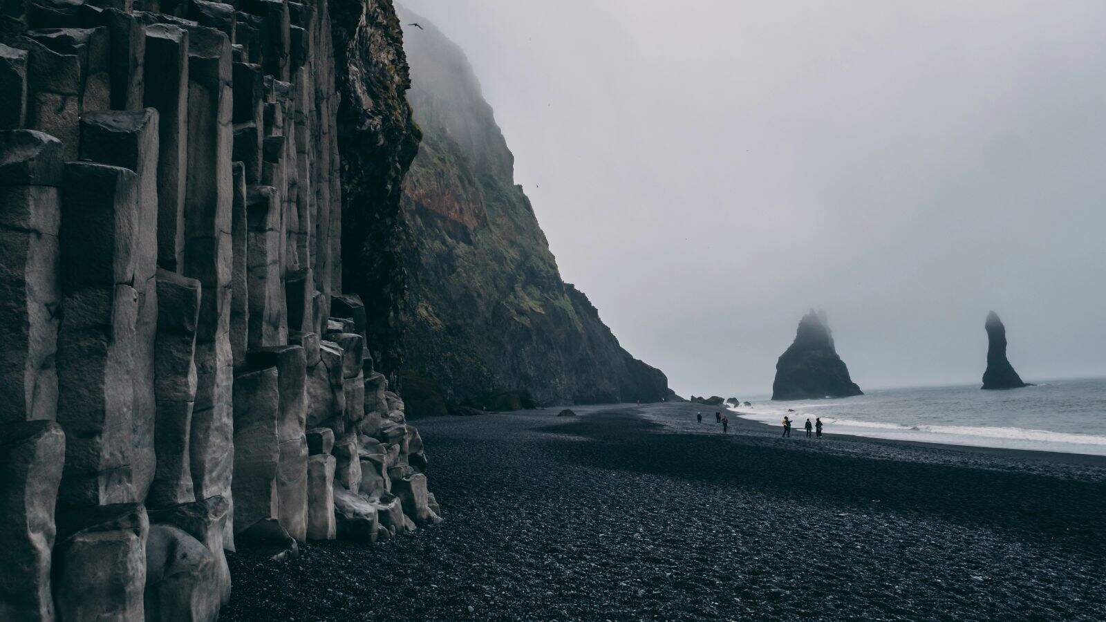 A black sand beach with geometric basalt columns on the left, misty cliffs in the background, and several sea stacks in the ocean; a few people walk along the shore under an overcast sky.