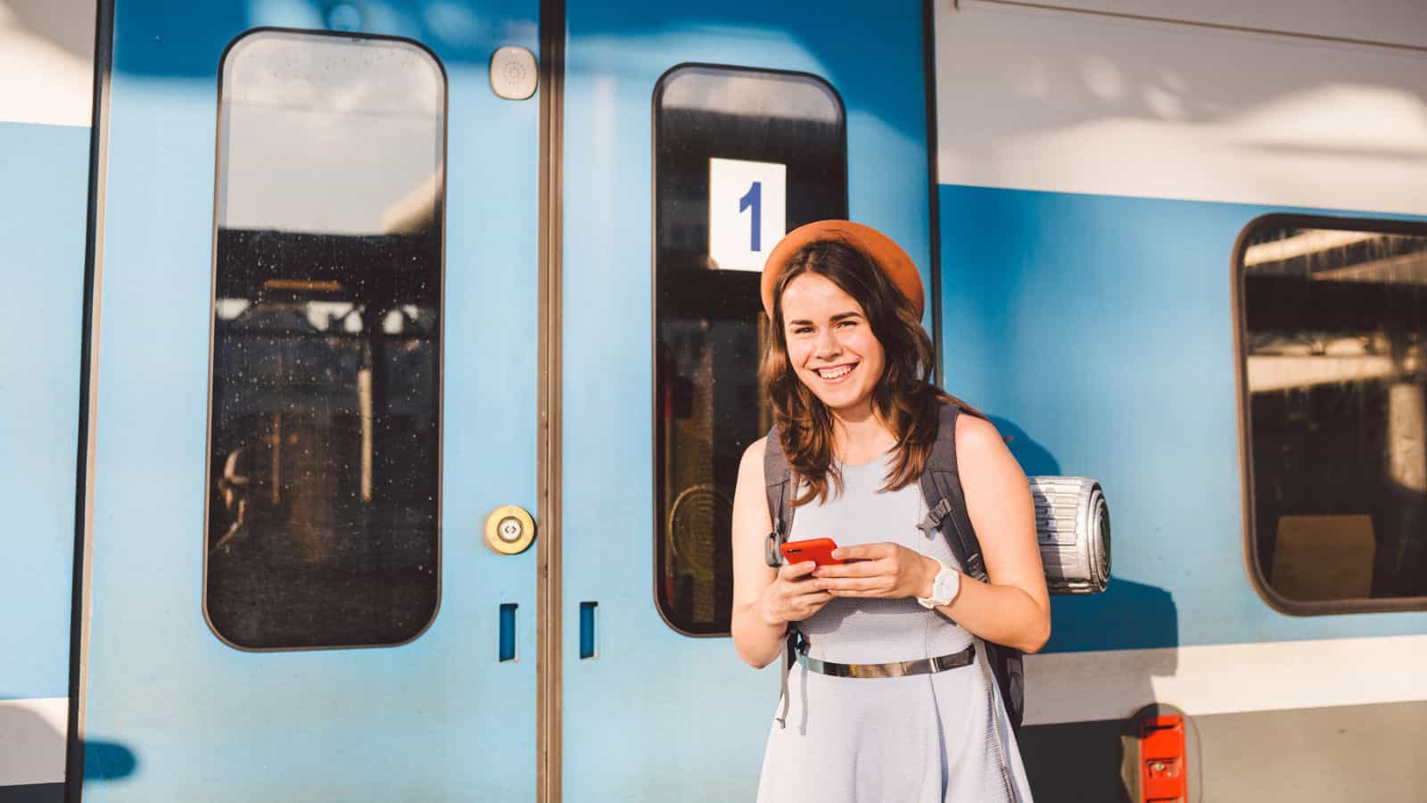A young woman wearing a hat and backpack stands in front of a blue train, smiling and holding a smartphone. She appears ready to board, and the train door has the number 1 displayed on it.