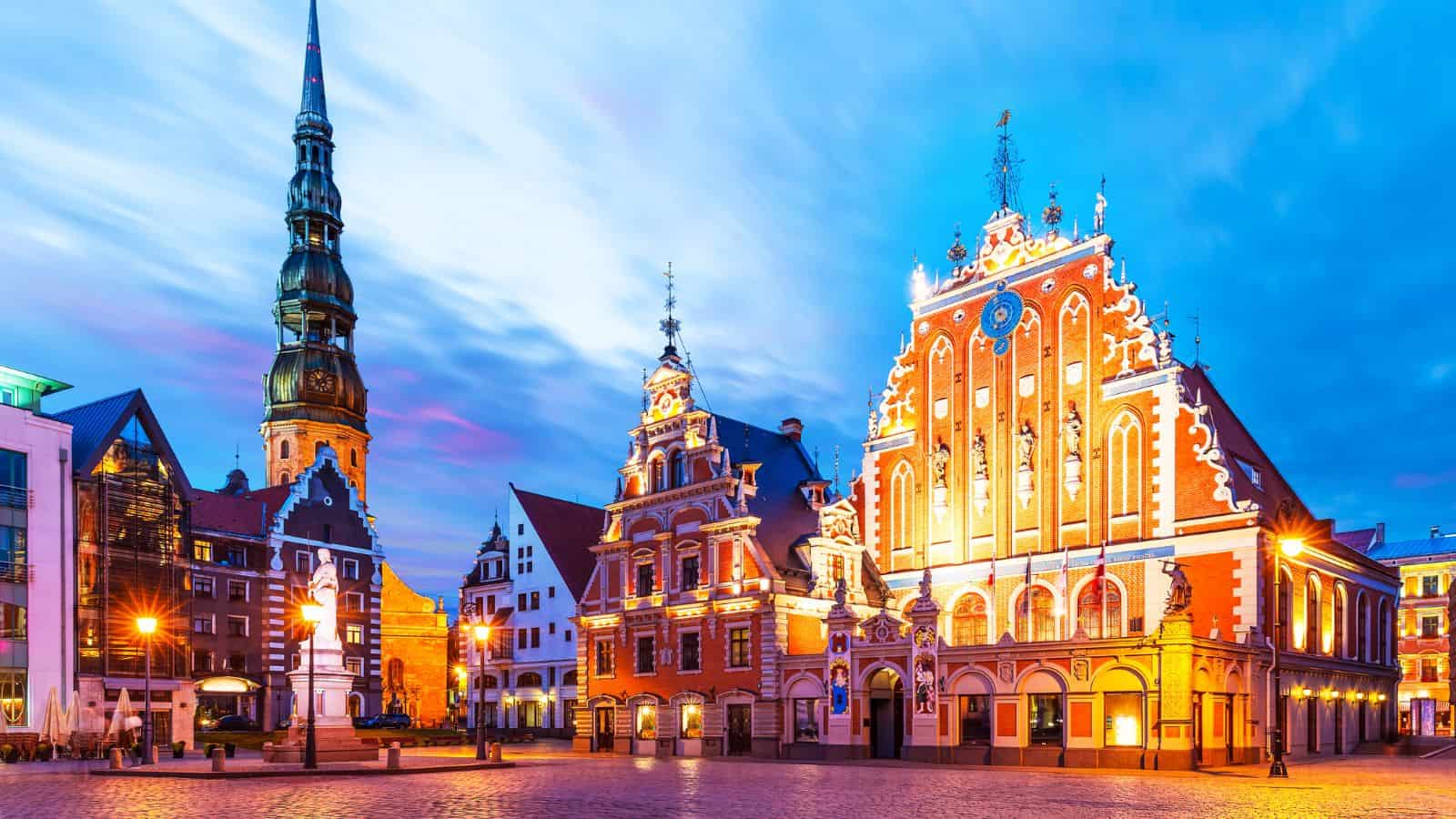 A city square at dusk features historic buildings with ornate facades and warm lighting. A tall church spire rises at left, and the sky displays blue and purple hues with clouds. The square is mostly empty.