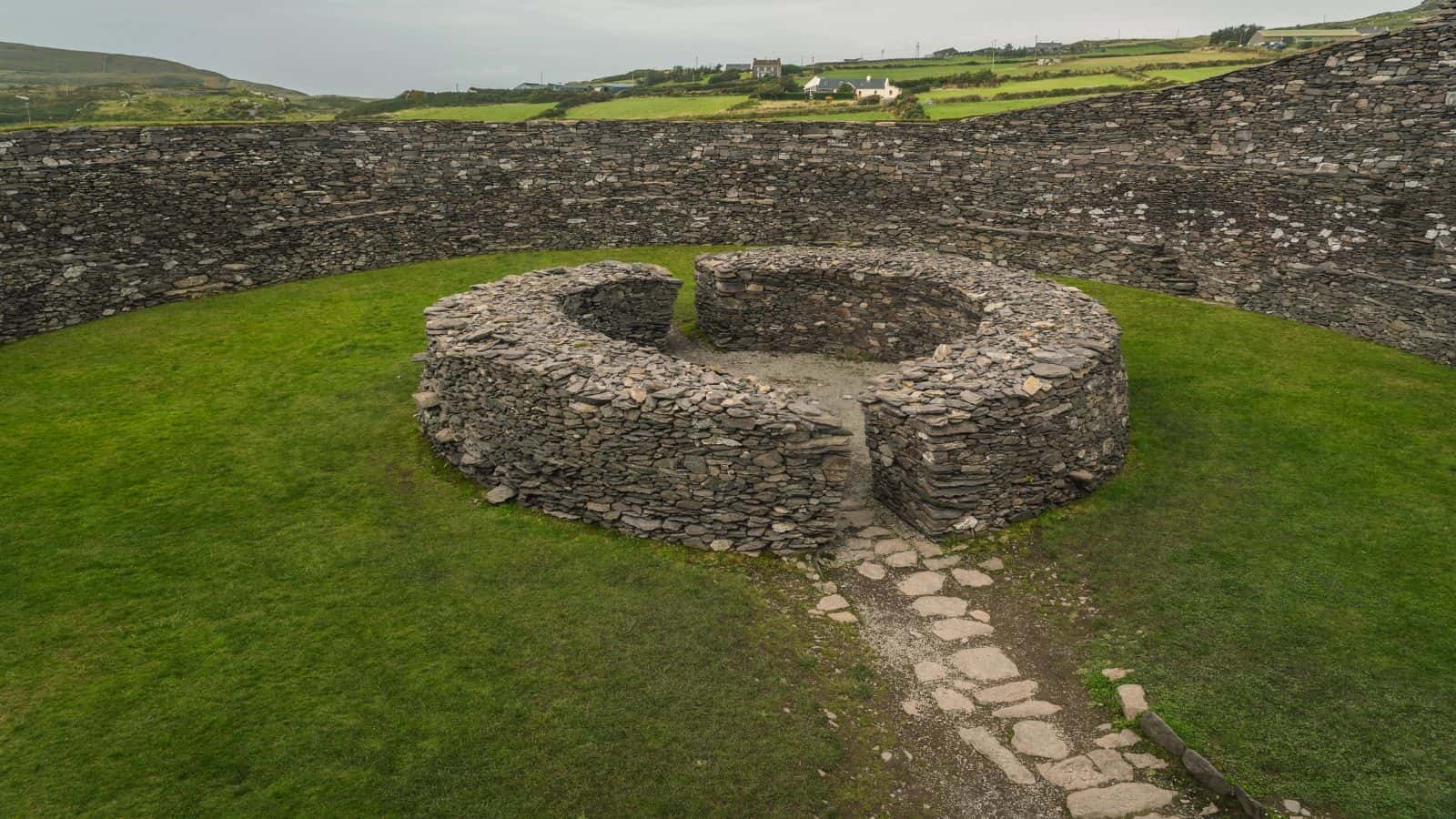 Ancient stone ringfort with circular inner wall on grassy field, surrounded by countryside and distant hills.