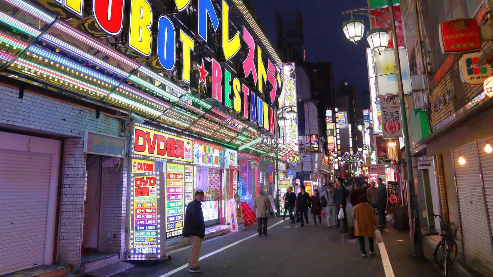 People walk along a brightly lit street with colorful neon signs in an urban area at night.