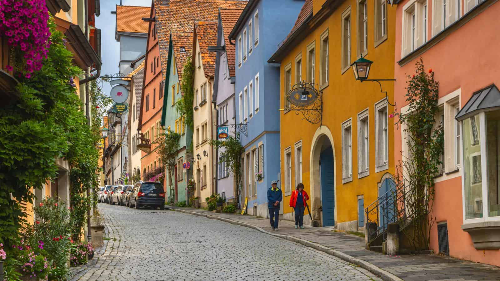 Colorful buildings line a cobblestone street where people walk and cars are parked in a quaint European town.