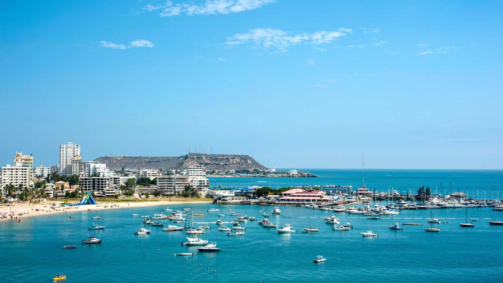 Boats anchored in a blue bay near a sandy beach with buildings and hills in the background under a clear sky.