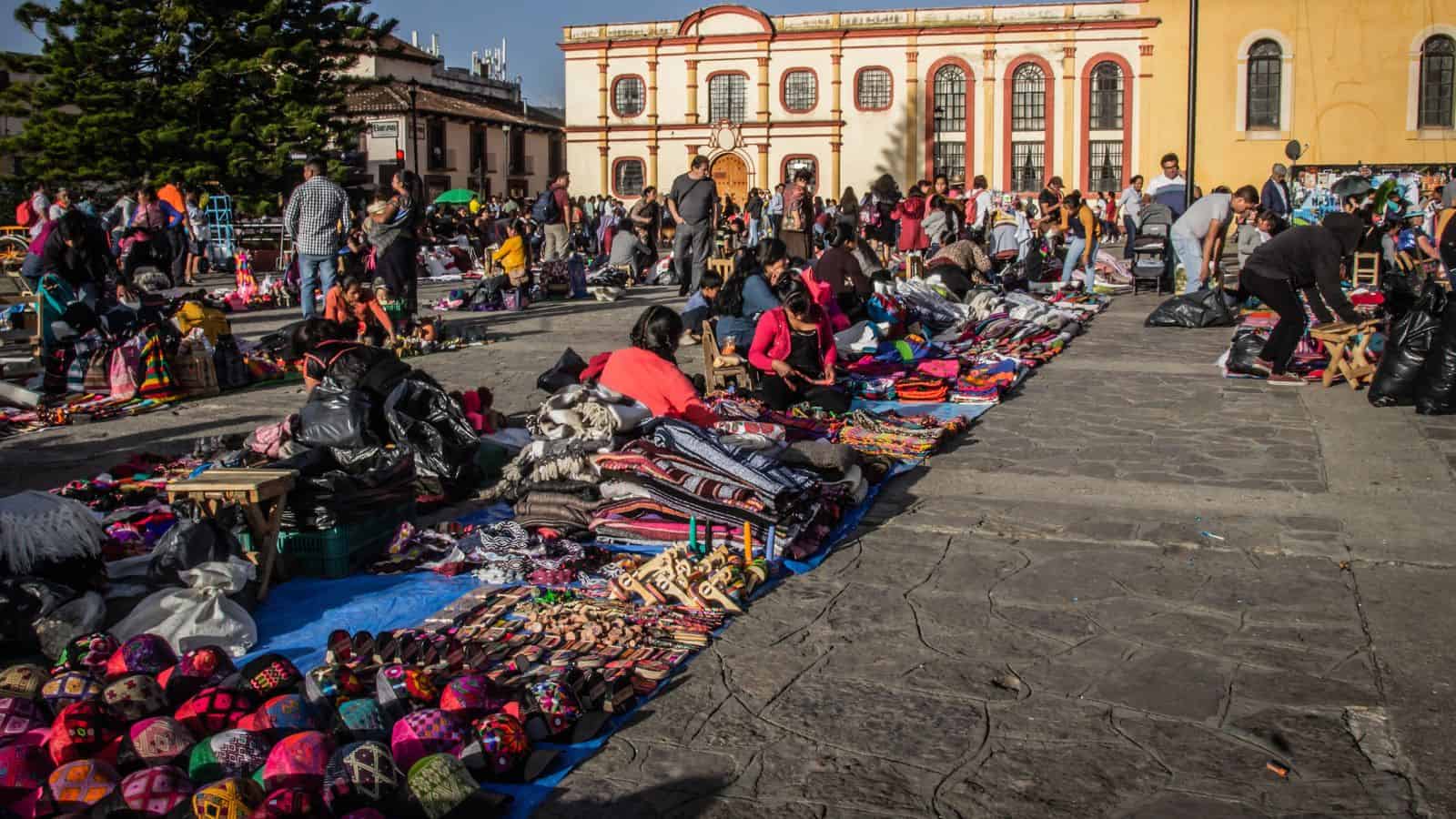 People shopping at an outdoor market with colorful textiles and crafts displayed on the ground in a sunny plaza.