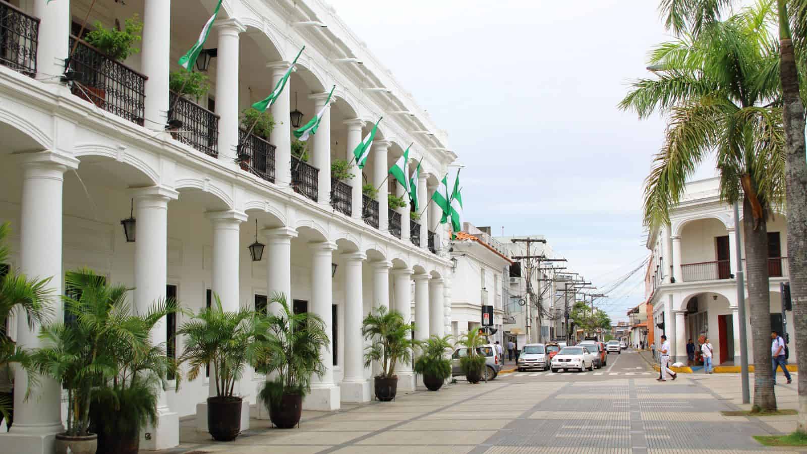 White colonial-style building with green-white flags, palm trees, and people walking on a city street.