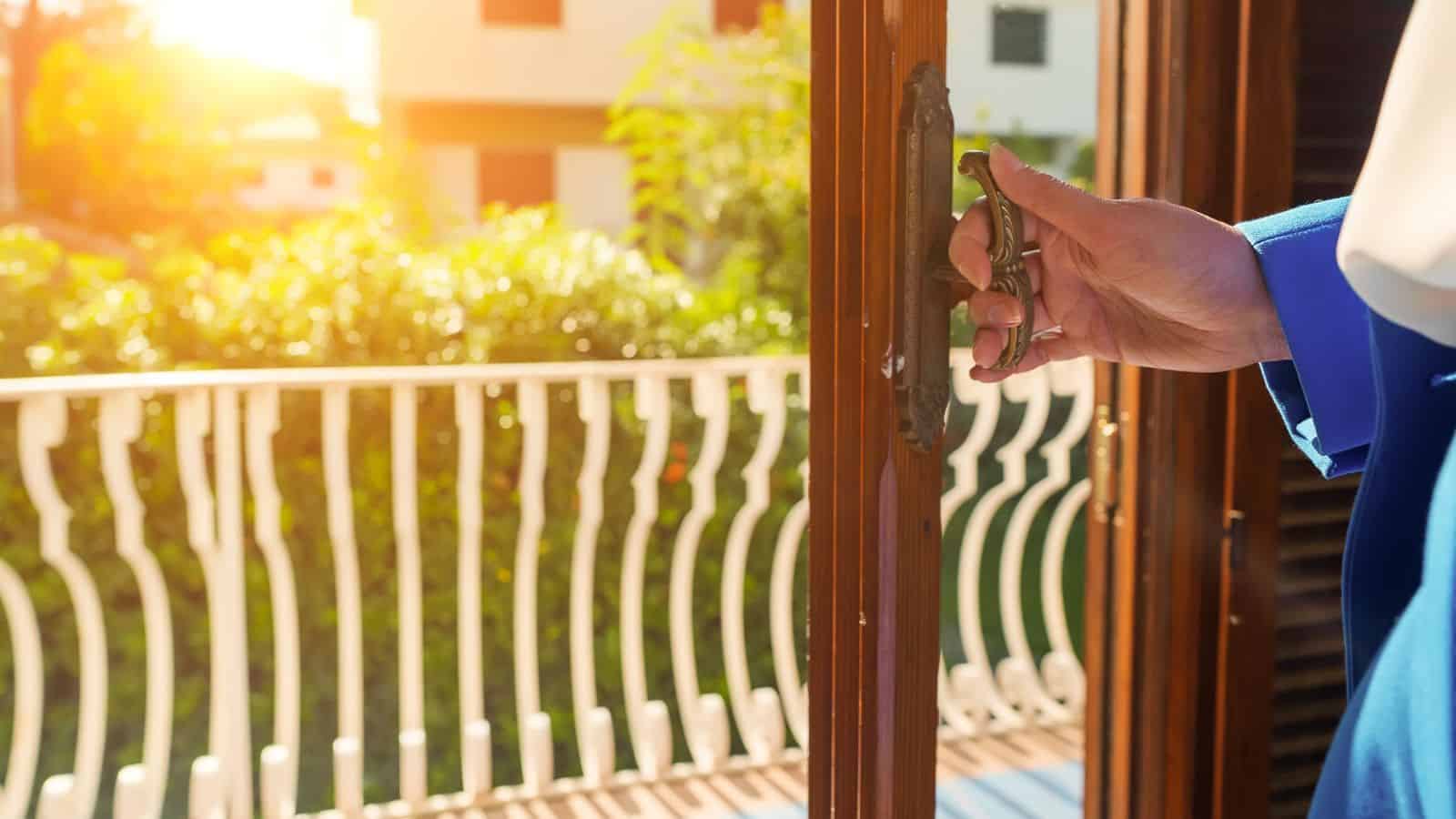 A person in a blue suit opens a glass door to a balcony with sunlight and greenery outside.