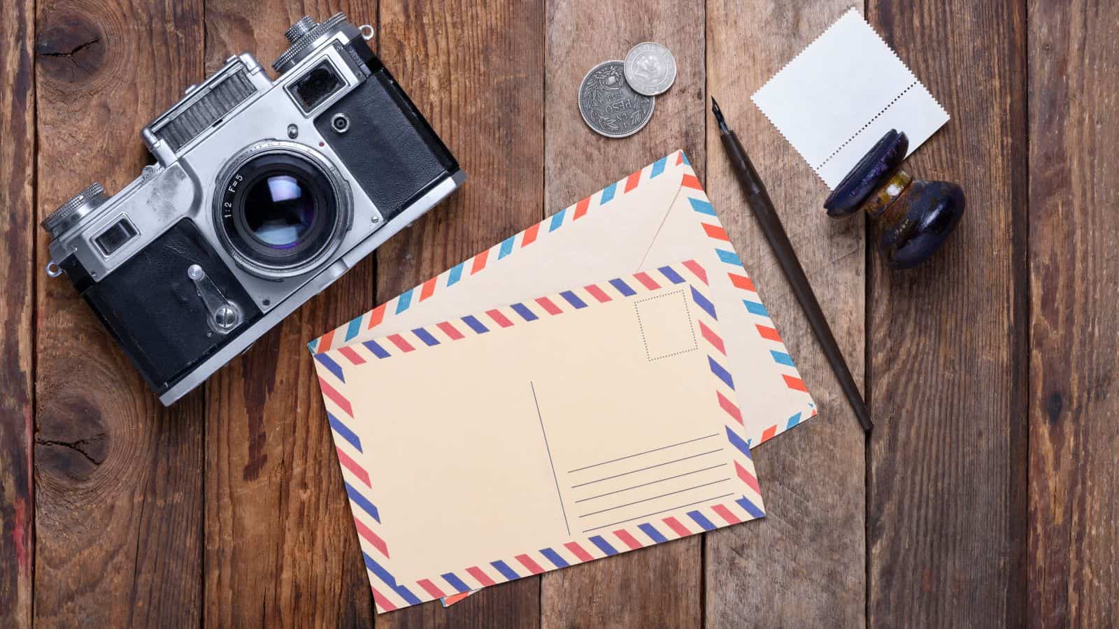 A vintage camera, two airmail envelopes, a fountain pen, two coins, a rubber stamp, and a blank postage stamp arranged on a wooden surface.
