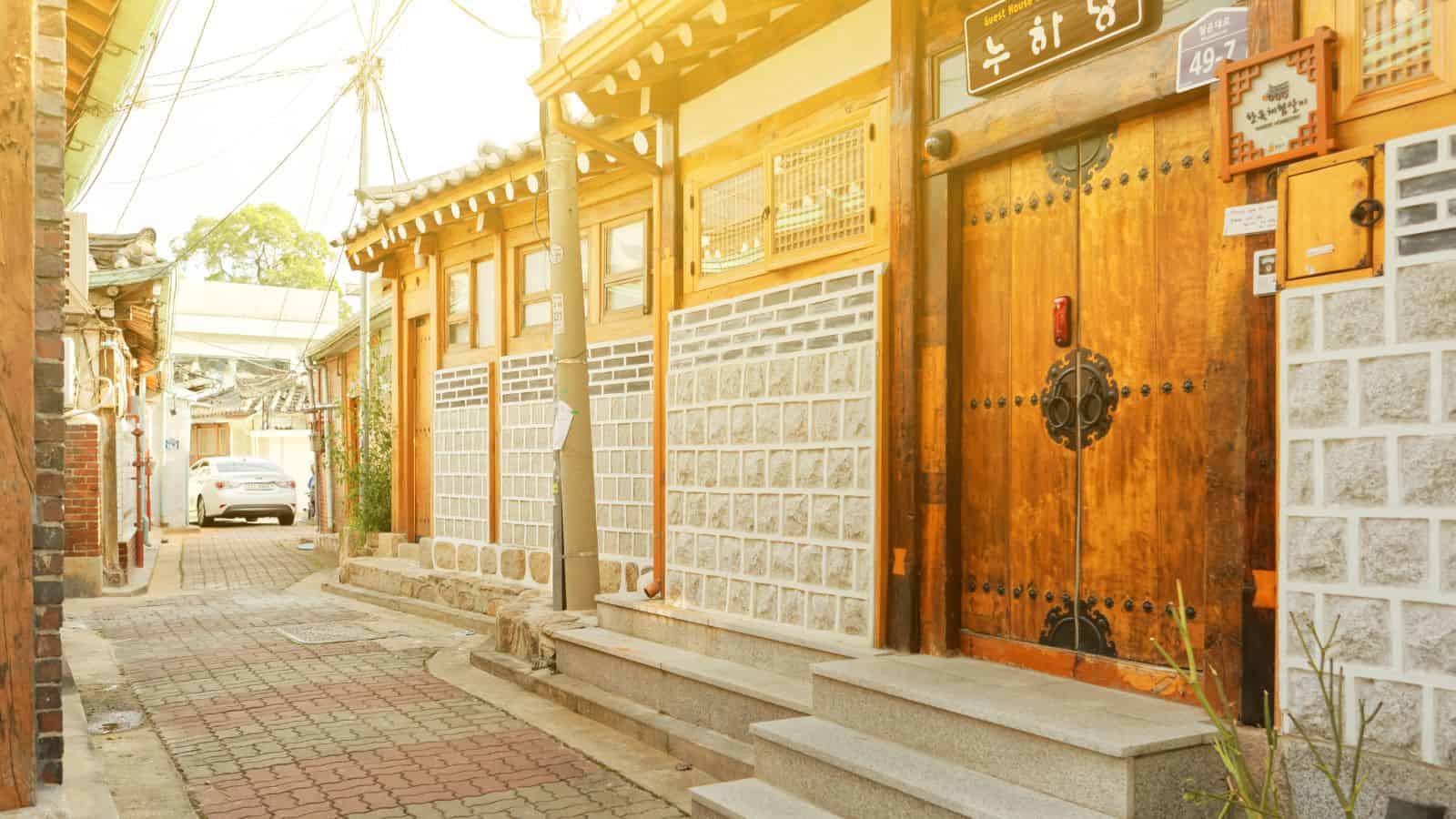 Narrow alley lined with traditional Korean hanok houses, wooden doors, stone walls, and sunlight filtering in.