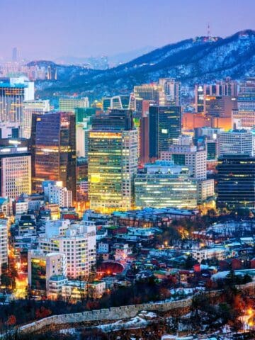 Cityscape of Seoul at dusk with illuminated high-rise buildings, residential areas, and Namsan Mountain in the background featuring the N Seoul Tower; snow is visible on the ground and rooftops—perfect for solo travel destinations.