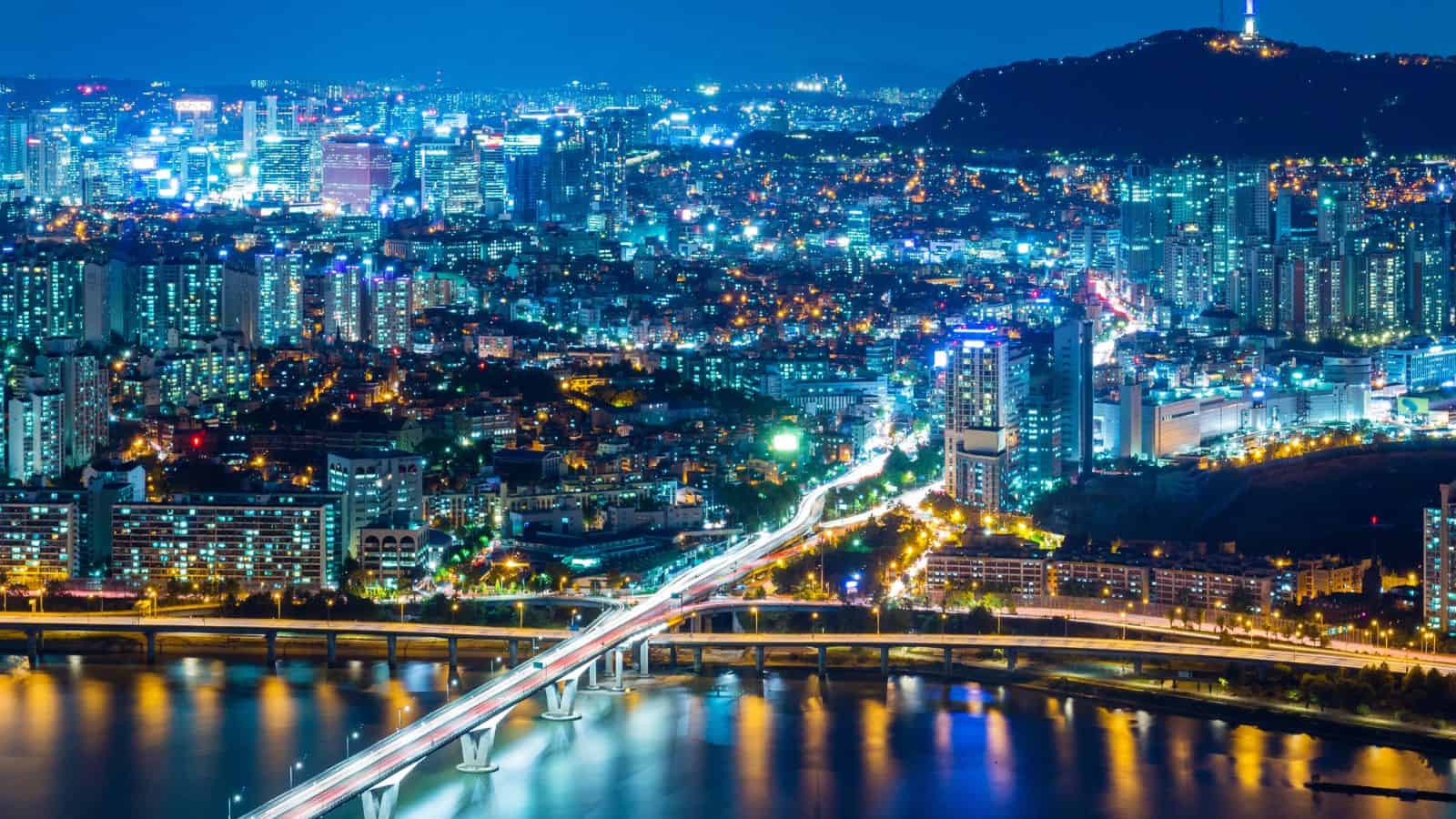 A brightly lit cityscape at night features a river with a bridge in the foreground, tall buildings with illuminated windows, and a hill with a lit tower—perfect for travelers seeking budget-friendly hotels nearby.