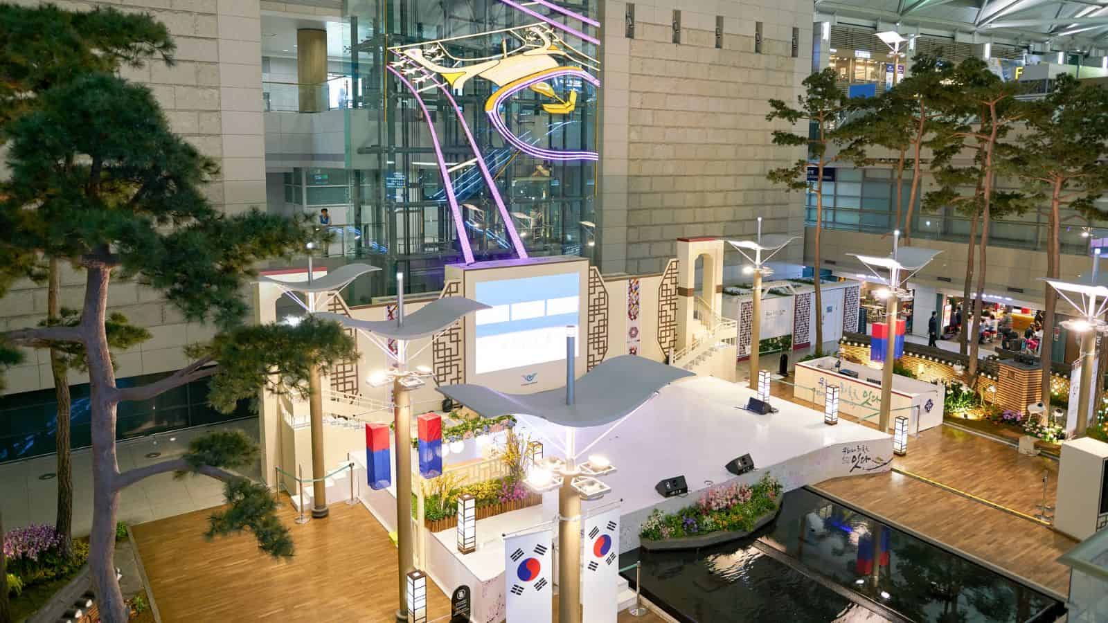 Indoor event space with a stage, large video screen, Korean flags, modern architecture, decorative lighting, and plants. The area has elevated walkways, glass windows, and a pond with reflections.