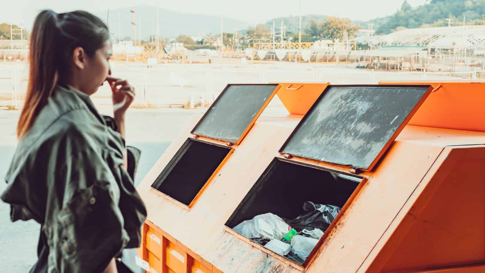 A person stands near an orange dumpster, looking thoughtfully at the garbage inside.