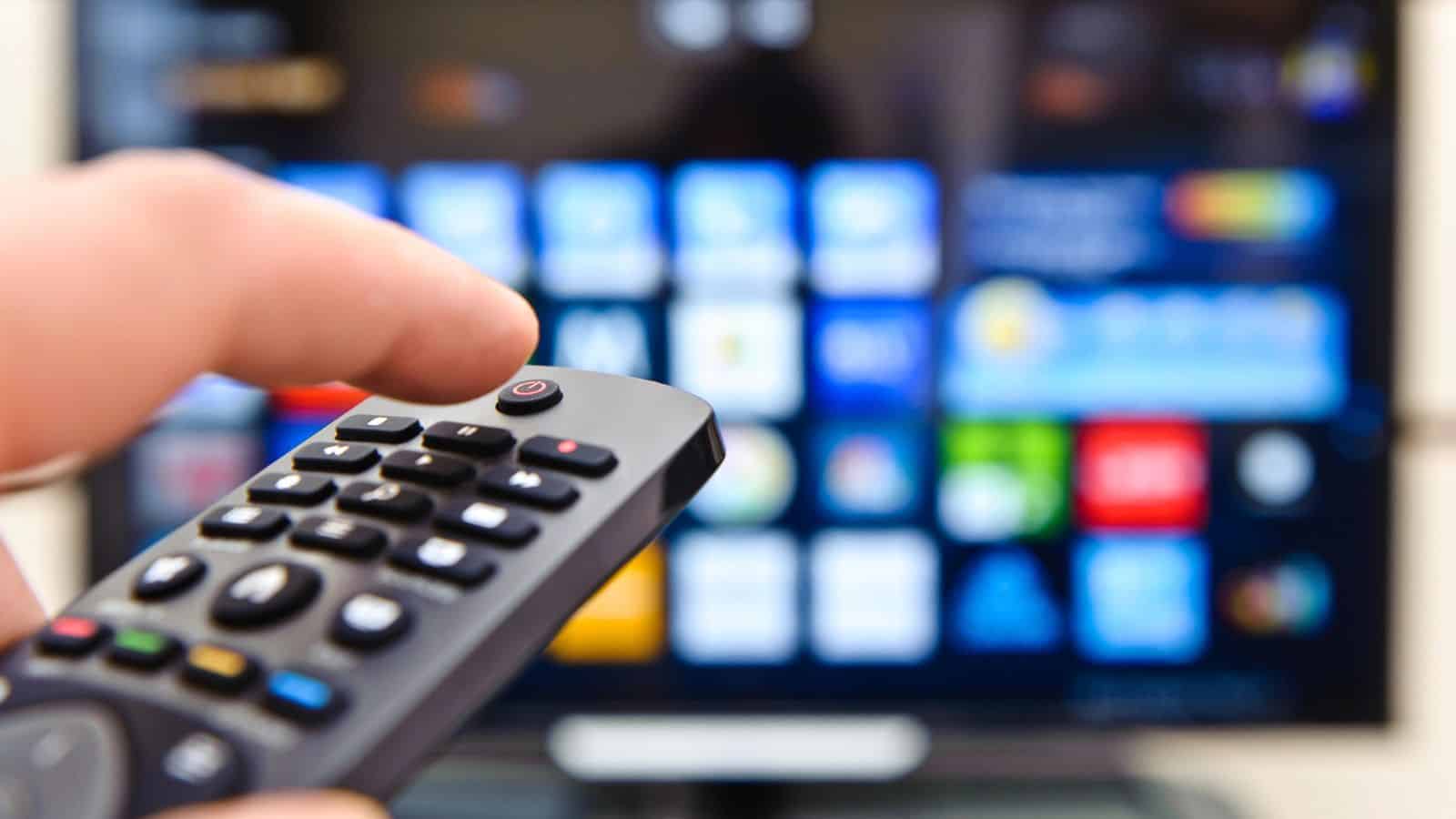 Hand holding a remote control pointed at a smart TV screen displaying app icons.