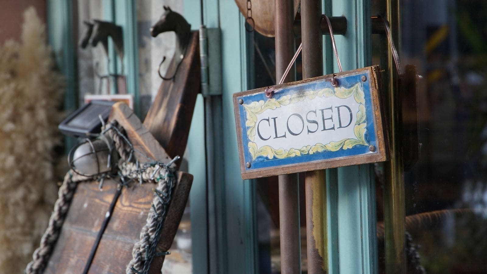 A "Closed" sign hangs on the handle of a glass door at a shop entrance. Various wooden items and decorations are visible beside the door—a common sight for first-time travelers noticing unique European habits.