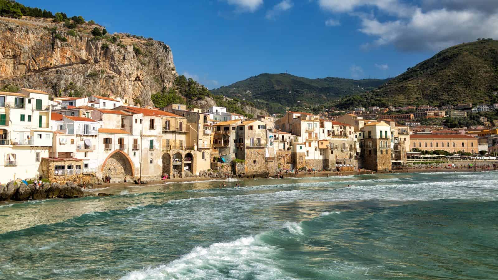Colorful seaside buildings line the shore with waves and mountains in the background under a partly cloudy sky.