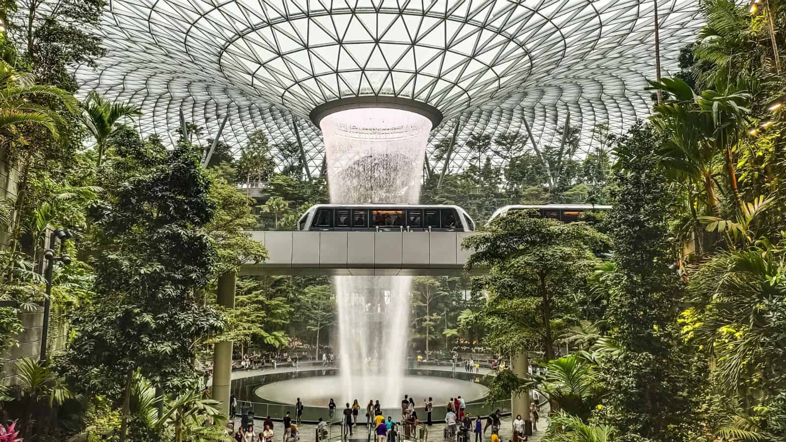 A wide indoor garden features a large waterfall at its center, with lush green trees surrounding it. A glass-domed roof covers the space, and a train runs across a bridge above the waterfall. People gather below.