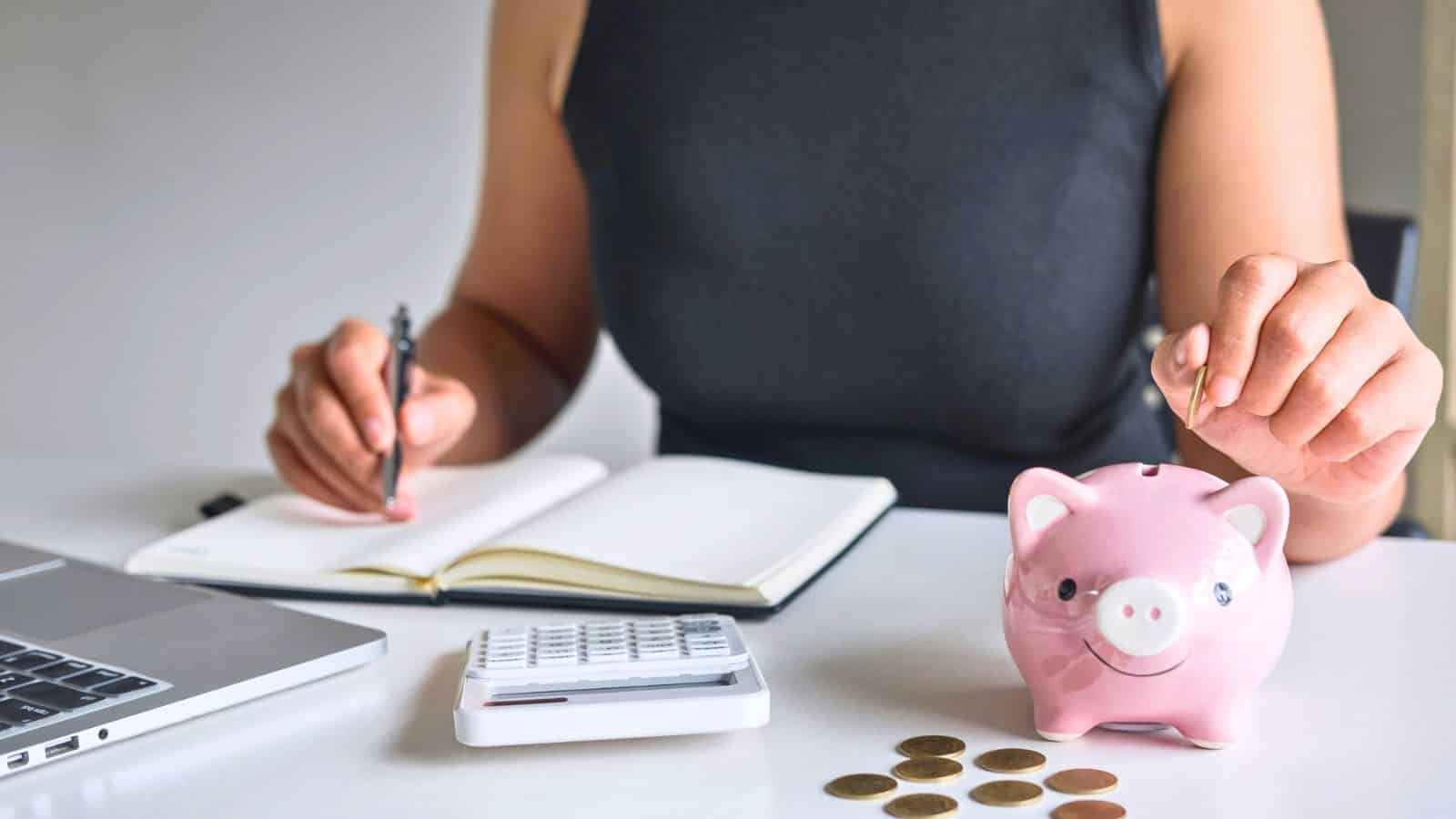 Person saving coins in a piggy bank, with a notebook, calculator, and laptop on the table.
