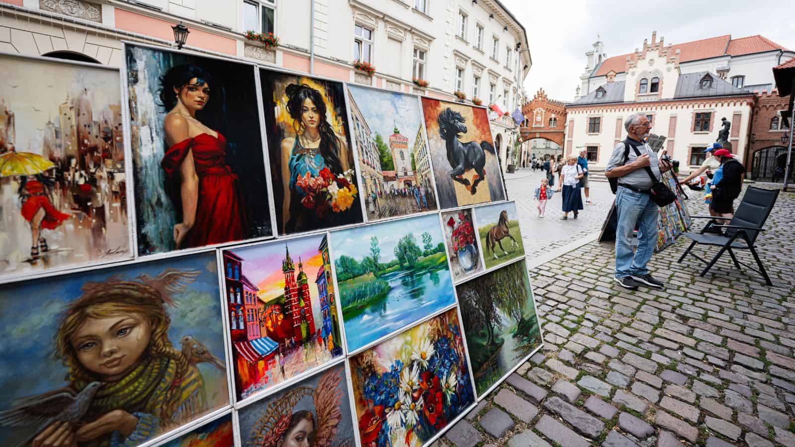 Outdoor art market with colorful paintings on display; people view the artwork along a cobblestone street.