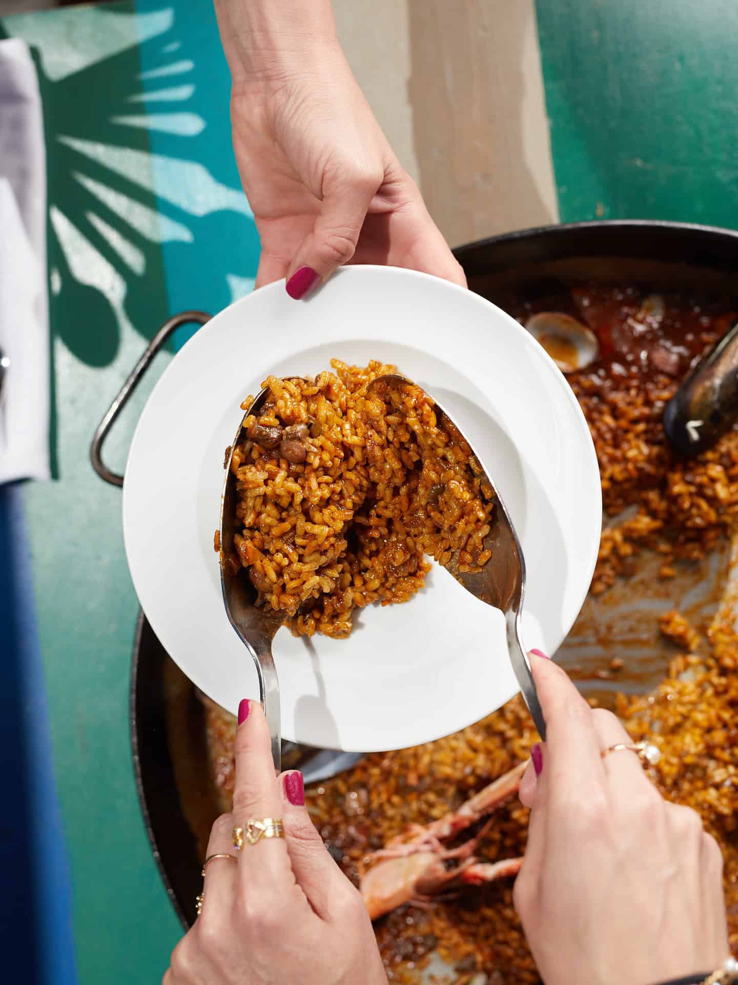 Two people serve a brown rice dish from a pan onto a white plate using a spoon and fork, enjoying the flavors reminiscent of the best restaurants Barcelona is known for.